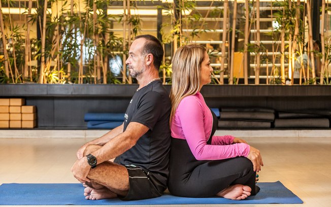 Para começar a yoga para casais, os dois sentam um de costas para o outro. Com isso, a dupla corrige a postura e ainda sente a respiração um do outro
