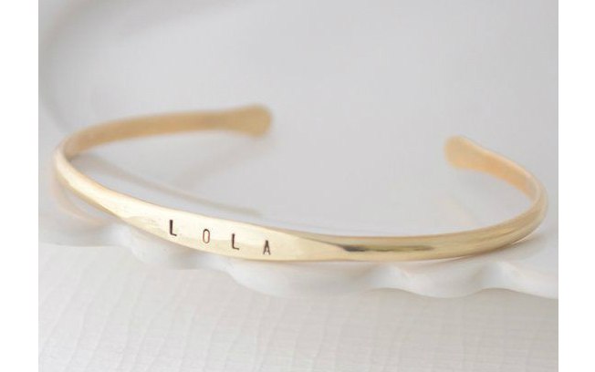 Finos braceletes dourados podem ter o nome do filho gravado. Fica discreto e elegante. De Etsy
